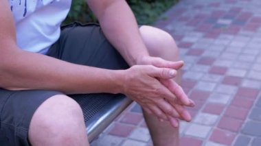 Male Rubbing Hands Sitting on a Bench in the Urban Park. A moment of waiting, excitement, anxiety, and emotional stress, trouble, nerves, and dread. Massage of hands, joints, fingers, skin. Outdoors.