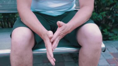 Male Rubbing Hands Sitting on a Bench in the Urban Park. A moment of waiting, excitement, anxiety, and emotional stress, trouble, nerves, and dread. Massage of hands, joints, fingers, skin. Outdoors.