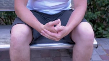 Male Rubbing Hands Sitting on a Bench in the Urban Park. A moment of waiting, excitement, anxiety, and emotional stress, trouble, nerves, and dread. Massage of hands, joints, fingers, skin. Outdoors.