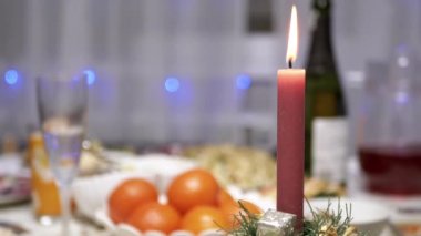 Burning Red Christmas Candle Burns with a Bright Flame on the New Years Table. Christmas decorated table on eve of holiday on background bulbs garlands. Cozy family dinner in a festive atmosphere.