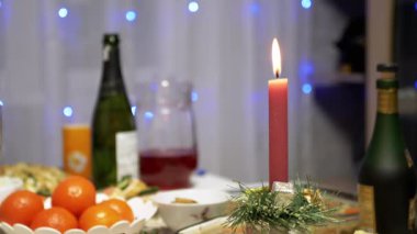 Burning Red Christmas Candle Burns with a Bright Flame on the New Years Table. Christmas decorated table on eve of holiday on background bulbs garlands. Cozy family dinner in a festive atmosphere.