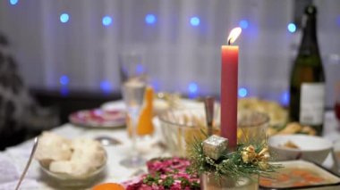 Burning Red Christmas Candle Burns with a Bright Flame on the New Years Table. Christmas decorated table on eve of holiday on background bulbs garlands. Cozy family dinner in a festive atmosphere.