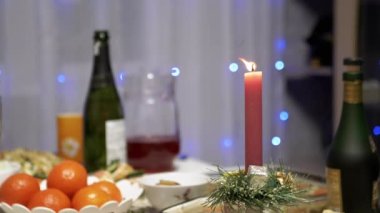 Burning Red Christmas Candle Burns with a Bright Flame on the New Years Table. Christmas decorated table on eve of holiday on background bulbs garlands. Cozy family dinner in a festive atmosphere.
