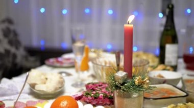 Burning Red Christmas Candle Burns with a Bright Flame on the New Years Table. Christmas decorated table on eve of holiday on background bulbs garlands. Cozy family dinner in a festive atmosphere.