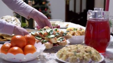 Kadın, yeni yıl arifesinde Noel Masası Servisi yapar. Eller masaya sandviç, portakal, abur cubur, salata, meyve suyu koyar. Bulanık Noel ağacı arka planı. Oturma odası. Evde aile yemeği..