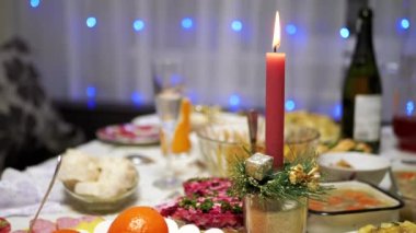 Burning Red Christmas Candle Burns with a Bright Flame on the New Years Table. Christmas decorated table on eve of holiday on background bulbs garlands. Cozy family dinner in a festive atmosphere.