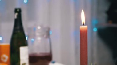 Burning Red Christmas Candle Burns with a Bright Flame on the New Years Table. Christmas decorated table on eve of holiday on background bulbs garlands. Cozy family dinner in a festive atmosphere.