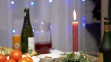 Burning Red Christmas Candle Burns with a Bright Flame on the New Years Table. Christmas decorated table on eve of holiday on background bulbs garlands. Cozy family dinner in a festive atmosphere.