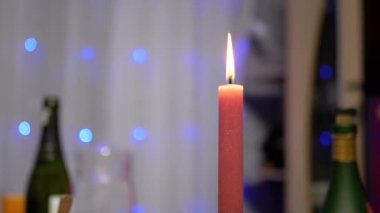 Burning Red Christmas Candle Burns with a Bright Flame on the New Years Table. Christmas decorated table on eve of holiday on background bulbs garlands. Cozy family dinner in a festive atmosphere.