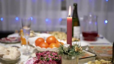 Burning Red Christmas Candle Burns with a Bright Flame on the New Years Table. Christmas decorated table on eve of holiday on background bulbs garlands. Cozy family dinner in a festive atmosphere.