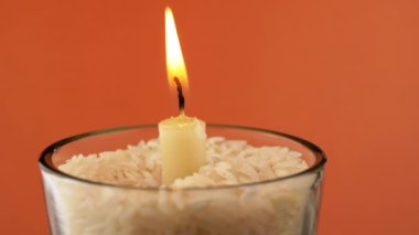 Rotating Burning Candle with a Bright Flame on an Isolated Orange Background. Candle in glass with rice. Concept of memory, ritual, celebration. Abstract, texture, colors. Slow-motion. Close-up.