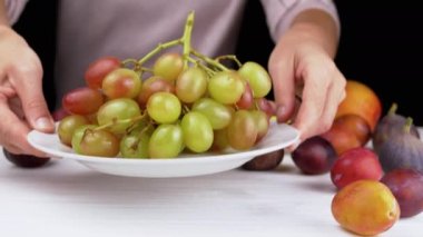 Eller masaya olgun üzümlü bir tabak koyar, tatmak için bir dut koparır. Bulanık meyve arka planında kırmızı ve yeşil renkli meyveler. Çiftçi kadın. Armutlar, erikler. Siyah beyaz. Hasat et.