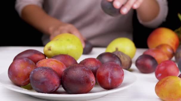 女性の手は プレートの上に置く熟した梅 ぼやけた背景果実に梨 皮の上に水が落ちるとジューシーな紫色のプラム 白いテーブルの上に果物がたくさんあります 秋の収穫 黒の背景 — ストック動画