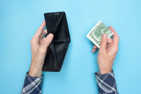 Young man holding an empty wallet and two dollars on a blue background. Financial literacy. Bankrupt. Money is tight. Importance of savings. Financial aid concept.
