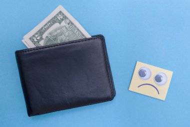 Cüzdanda iki dolar var. Hüzünlü ifade cüzdana bakar. Mavi arka plan. İflas etti. Para sıkıntısı var. Birikimlerin önemi. Mali yardım kavramı.