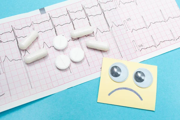 EKG Elektrokardiyogram, haplar ve üzgün gülümseme. Kalp kardiyogramda yatıyor. Kalbin sağlığıyla ilgilenmek. Mavi arka plan. Tıbbi araştırma. Yakın plan.
