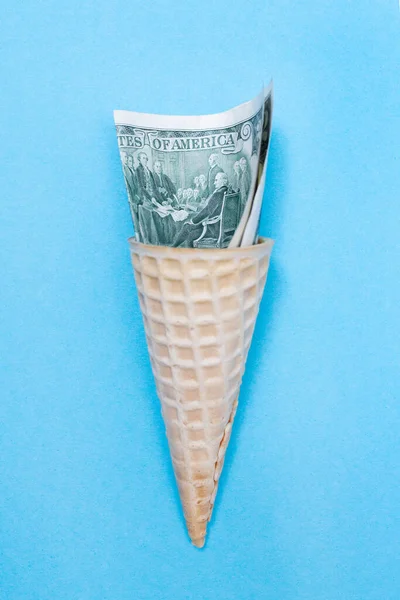 Ice cream cone and American US dollars on a blue background. Waffle cone. Finance. Inflation. Money is tight. Vertical. Flat lay Стоковая Картинка