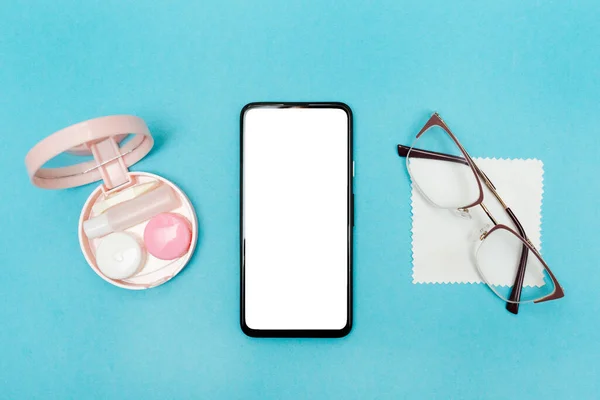 Mavi arka planda kontak lensler için gözlük ve seyahat seti. Beyaz ekranlı telefon. Gözlükle lens arasında seçim yapmak. Vizyon sağlığıyla ilgilenmek. Modelleme. Boşluğu kopyala.