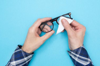 Gözlükleri peçeteyle silen bir adam. Temizlik bardakları. Mavi arka plan. Düz yatıyordu. Üst görünüm.