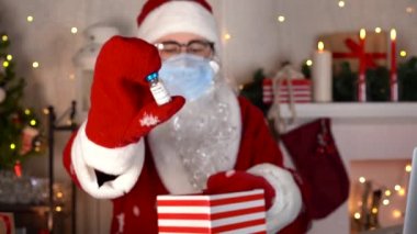 Mavi maskeli Noel Baba, Covid aşı şişesini hediye kutusuna koyar..