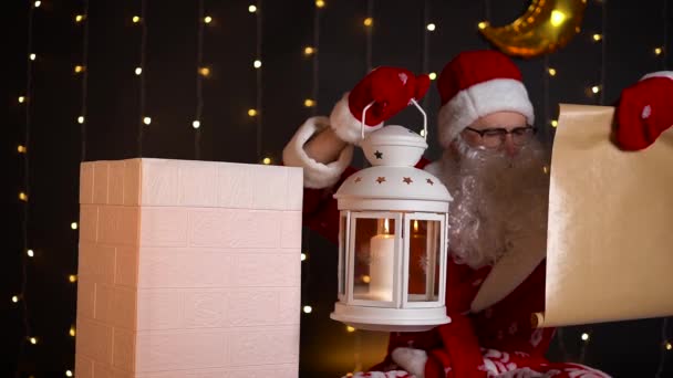 Santa checking wish list with candle lamp near house chimney at Christmas night. — Stock Video