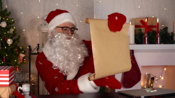 Père Noël traditionnel vérifier liste de souhaits avec des boîtes-cadeaux près d'une cheminée confortable. — Video