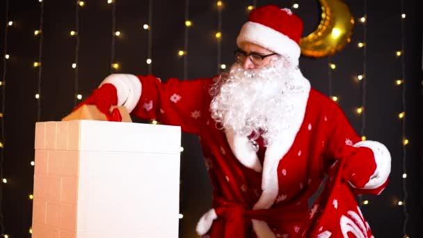 Санта Клаус достает рождественские подарки из мешка и кладет их в дымоход — стоковое видео