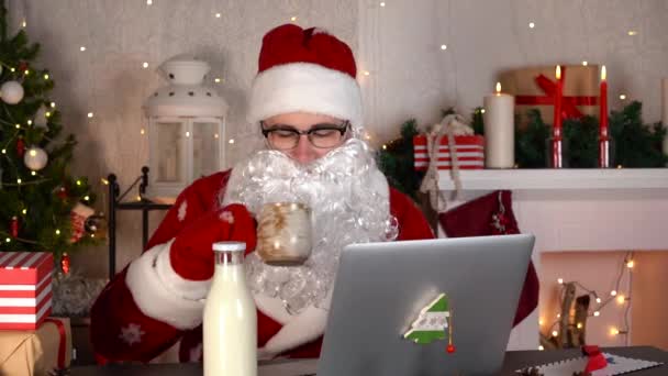 Der Weihnachtsmann in seiner Weihnachtswerkstatt mit Laptop und leckerer Milch. — Stockvideo