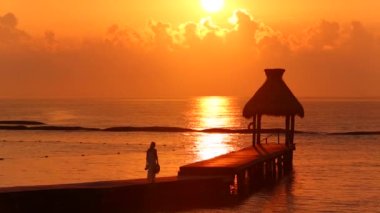 Kadın gün batımında tropik tatil köyünde rıhtımda yürüyor.