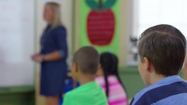 Öğretmen sınıfta ders veriyor ve öğrenciler el kaldırıyor, çocuk ellerine odaklanıyor.