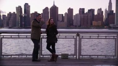 New York 'ta bir çift nehir kenarında durup, arka planda ufuk çizgisi ile konuşuyor.