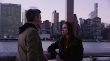 New York 'ta bir çift nehir kenarında durup, arka planda ufuk çizgisi ile konuşuyor.