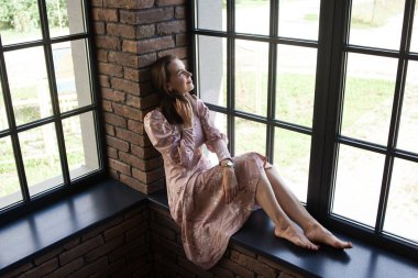 Pembe elbiseli güzel bir kadın pencerenin kenarında çıplak ayakla oturuyor.