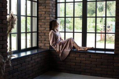 Pembe elbiseli güzel bir kadın pencerenin kenarında çıplak ayakla oturuyor.