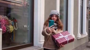 Kahverengi ceketli kız hediye kutusuyla selfie çekiyor.