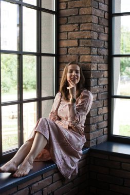 Pembe elbiseli güzel bir kadın pencerenin kenarında çıplak ayakla oturuyor.