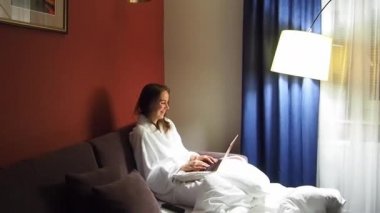 Bornoz giymiş bir otel odasında dizüstü bilgisayar kullanan bir iş kadını.