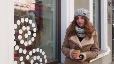 Kahverengi paltolu kız elinde kahve ile caddede yürüyor.