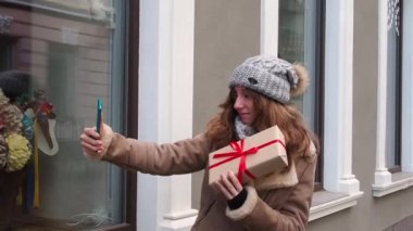 Kahverengi ceketli kız hediye kutusuyla video görüşmesi yapıyor.