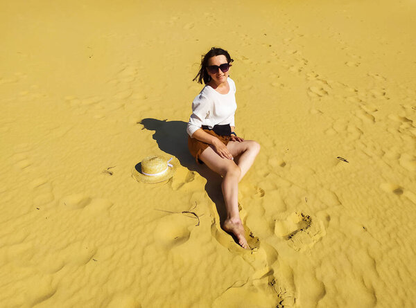 Молодая женщина в белой блузке и соломенной шляпе сидит в пустыне под голубым небом. Девушка сидит в песчаных дюнах в украинской пустыне, природный парк "Олешские пески", Олешковский писки, Херсонская область