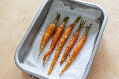Glazed and roasted young red carrots on a piece of baking paper, baked baby carrots on the wooden background. Roasted young whole carrot with greens and sea salt in baking tray.