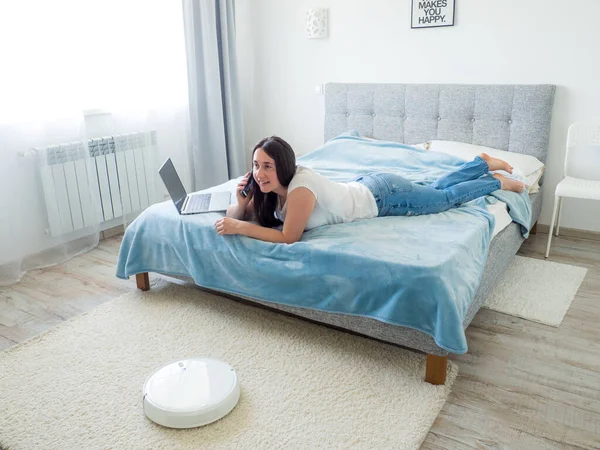 White robotic vacuum cleaner cleaning the floor while woman sitting on the bed and using phone. Smart technology concept. Woman in white t-shirt and blue jeans enjoying home cleaning. Work at home.