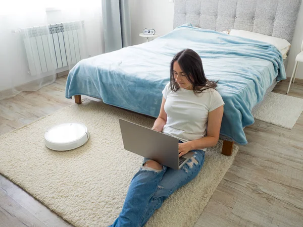 White robotic vacuum cleaner cleaning the floor while woman sitting near bed and using laptop. Smart technology concept. Woman in white t-shirt and blue jeans enjoying home cleaning. Work at home.