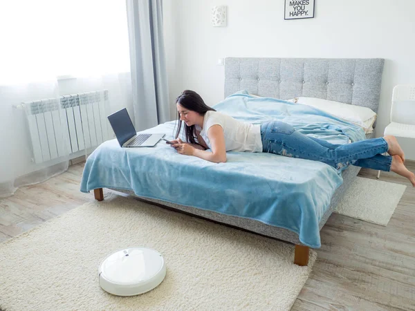 White robotic vacuum cleaner cleaning the floor while woman sitting on the bed and using phone. Smart technology concept. Woman in white t-shirt and blue jeans enjoying home cleaning. Work at home.