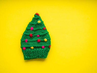 Green knitted hat in the form of a Christmas tree on a yellow background. Christmas, warming and handmade concept. New year greeting card with copy space. Knit for children.
