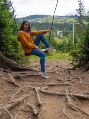 Woman with dark hair in a mustard sweater sitting on a swing and mountain view. Calm and quiet wanderlust concept moment when person feels happiness and life. Alone travel in Ukraine.