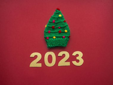 2023 and green knitted hat in the form of a Christmas tree on a red background. Christmas, warming and handmade concept. New year greeting card with copy space. Knit for children.
