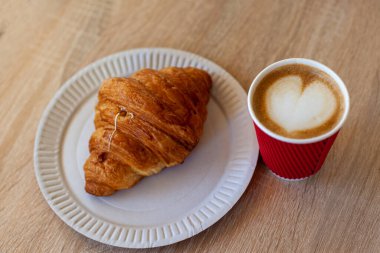 Cup of cappuccino with latte art and croissant on wooden table. Beautiful foam, red paper cup. Pastry and Coffee to go on wooden background with heart shaped latte art on top. Morning breakfast. 