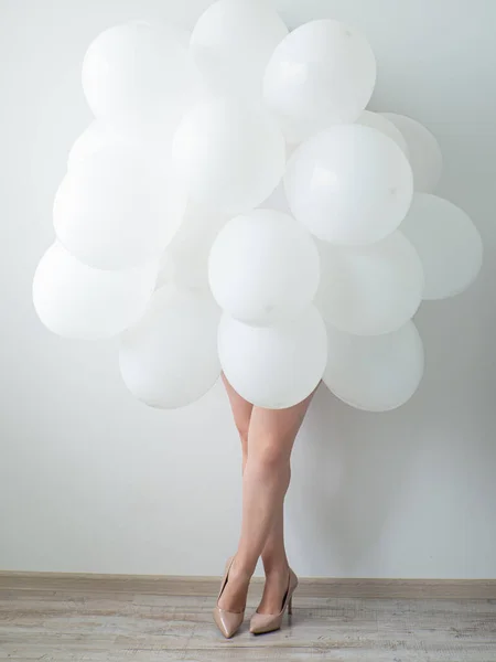 Woman in beige high heels holding white color balloons on a white wall background. Legs and a lot of white air balloons. Happy birthday anniversary. White decoration. Copy space