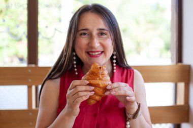 Young beautiful woman in red blouse eating croissant at a cafe. Hungry female having breakfast out while eating tasty croissant with pleasure.